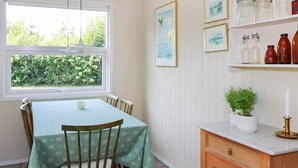 A dining area with a table and chairs, a window with a view of greenery, and a shelf with jars and a candle.