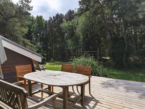 A wooden deck with a round table and chairs, surrounded by a forested area.