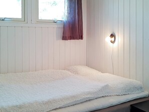 Et soveværelse med en hvid madras, et vindue med gardin og en vægmonteret lampe.