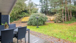 A wooden deck with outdoor furniture, surrounded by a garden with trees and a hedge. - 6 Person Holiday Home in Ansager (Ansager)