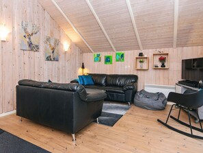 A cozy living room with a black leather sofa, a grey bean bag, and a black rocking chair. There are wooden walls, a wooden ceiling, and a TV on a wooden stand.