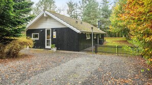 A small, dark wooden cabin with a white door and windows, surrounded by gravel and a wooden fence, set in a wooded area. - 6 Person Holiday Home in Ansager (Ansager)