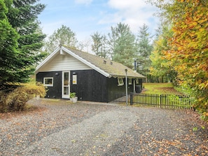 A small, dark wooden cabin with a white door and windows, surrounded by gravel and a wooden fence, set in a wooded area. - 6 Person Holiday Home in Ansager (Ansager)