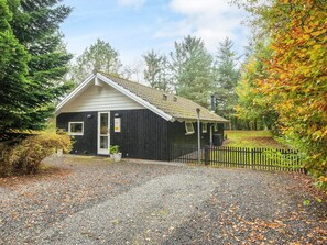 A small, single-story building with a sloped roof, a gravel driveway, and a wooden fence.
