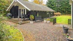 En liten, mörk trästuga med ett tak täckt av tegelpannor, en veranda och en inhägnad trädgårdsdel.