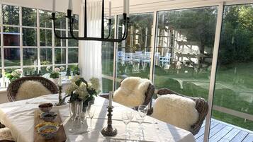A dining area with a table set for six, surrounded by wicker chairs, a chandelier, and a view of a garden through large windows.