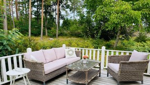 Eine Holzterrasse mit Korbmöbeln, darunter eine Sitzecke mit Sofa und zwei Sesseln, sowie ein kleiner runder Tisch.