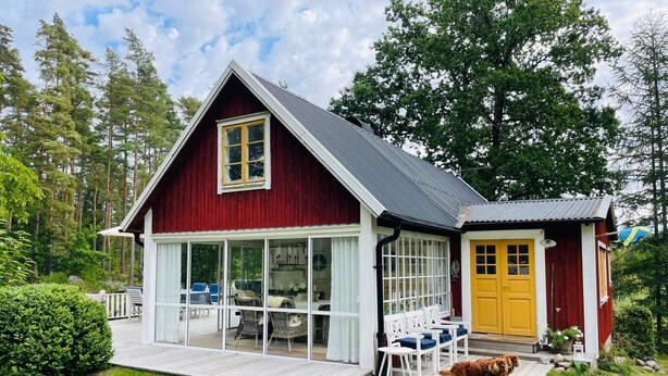 Ein rotes Holzhaus mit einer gelben Tür, einer Veranda mit weißen Stühlen und einer kleinen Holzterrasse.