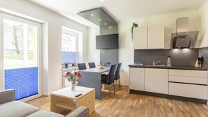 Una sala de estar moderna con sofá, una mesa de comedor con sillas, una zona de cocina con armarios y un microondas, y un ventanal con persianas.