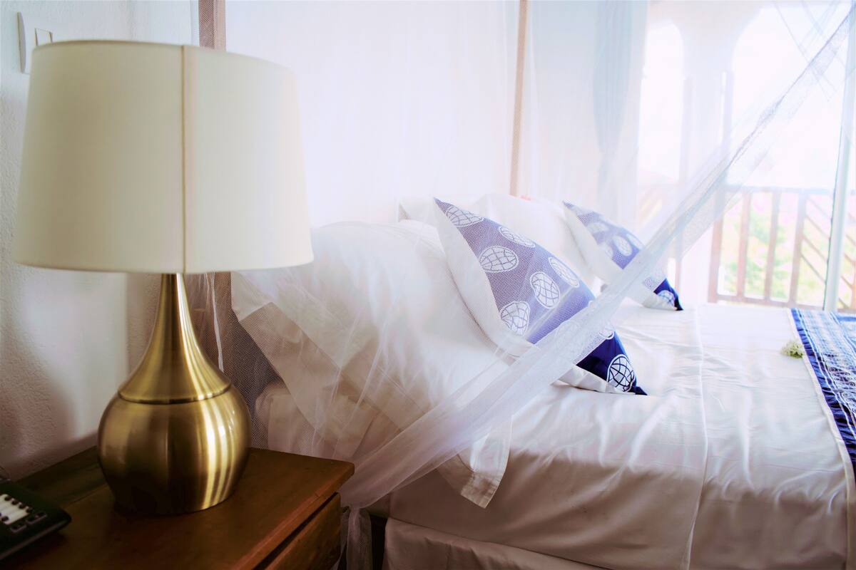 Cama bem arrumada com lençóis brancos e almofadas estampadas em azul, uma luminária dourada sobre uma mesa de cabeceira de madeira e uma cortina translúcida parcialmente aberta.