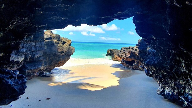 Vista de una playa a través de la entrada de una cueva, con agua azul cristalina y una orilla arenosa.