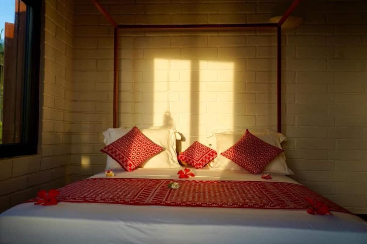 Cama com roupa de cama branca e almofadas vermelhas estampadas, diante de uma parede de tijolos com uma janela que ilumina o espaço.