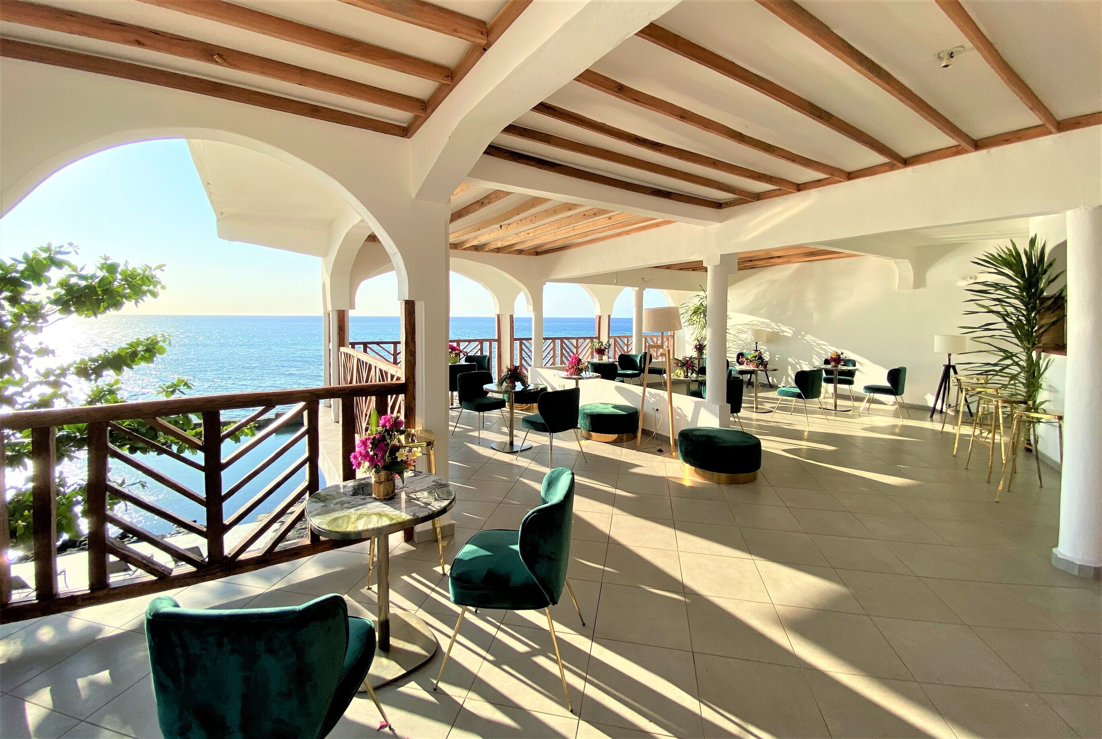 A spacious indoor area with a view of the sea, featuring wooden beams, green chairs, and a dining table.
