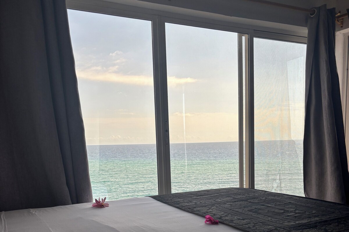 A hotel room with a large bed, a view of the sea through floor-to-ceiling windows, and a dark-colored rug.