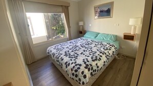 A bedroom with a bed, a ceiling fan, a window with curtains, a nightstand with a lamp, and a wall-mounted picture.