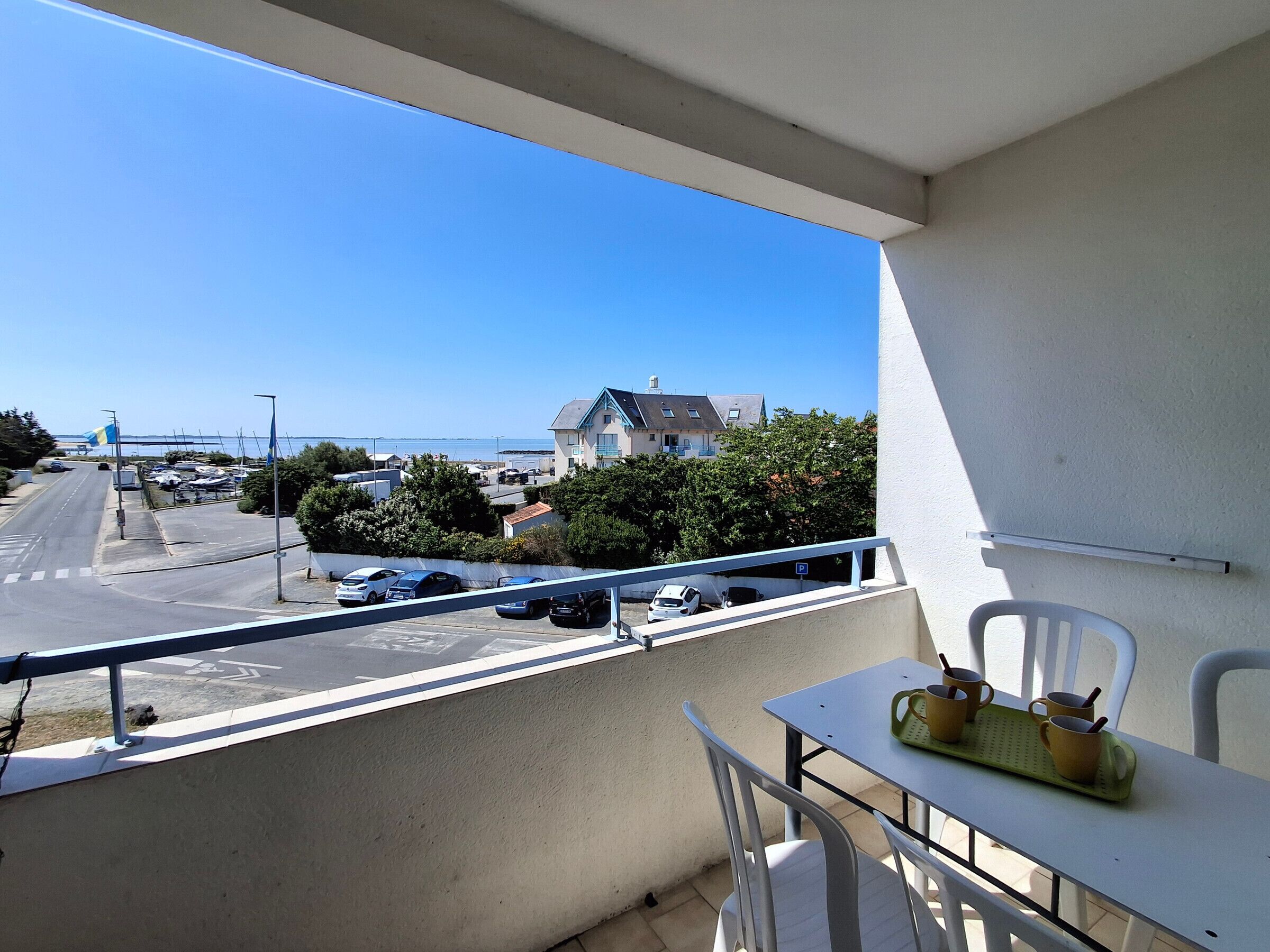 A balcony with a table and chairs, overlooking a street with parked cars and a building with a blue roof.
