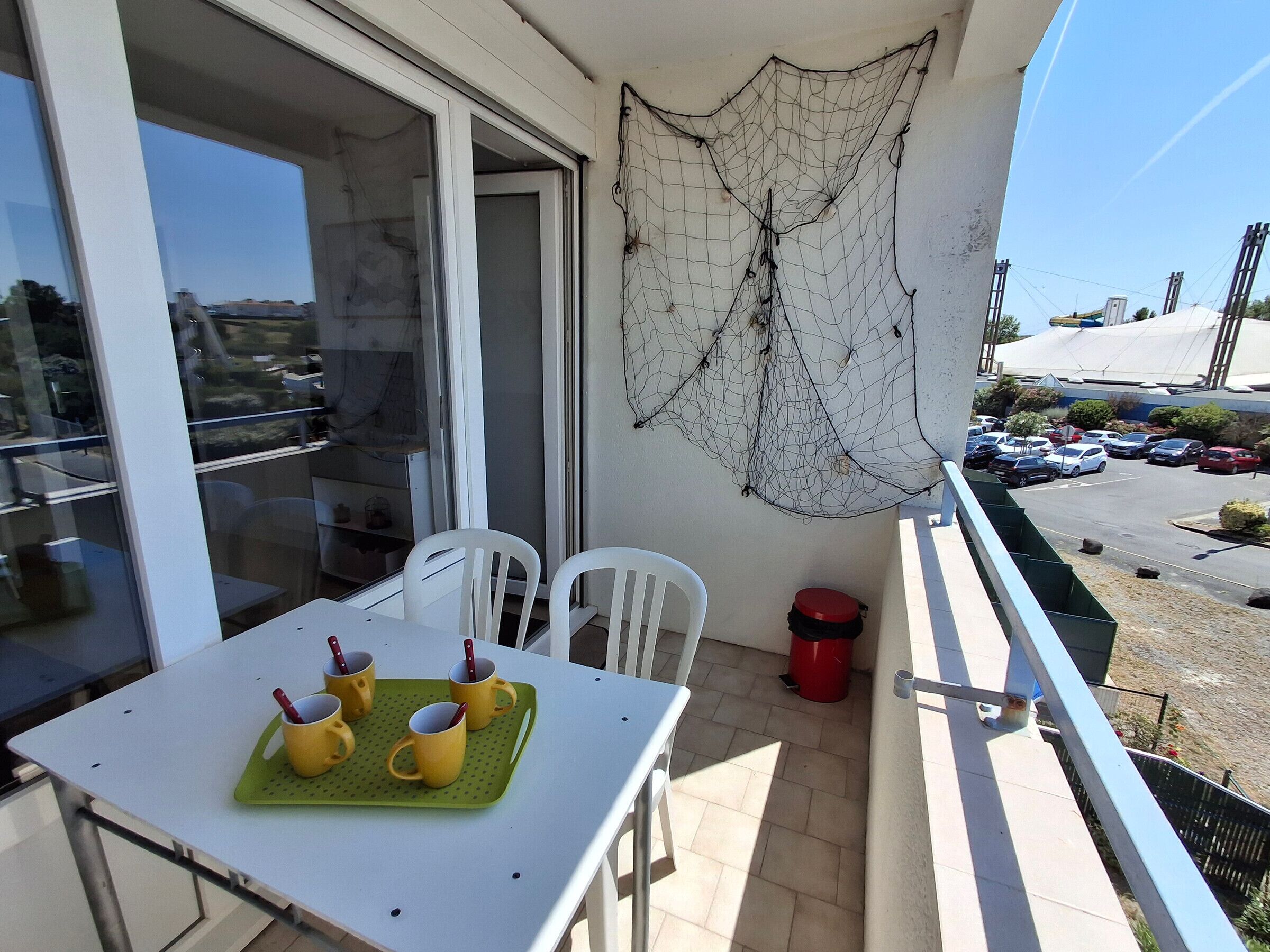 Ein Balkon mit Tisch, gelben Tassen, einer grünen Tablett, weißen Stühlen und einem roten Mülleimer.