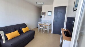 Ein kompakter Wohnraum mit einem dunkelgrauen Sofa, zwei gelben Kissen, einem weißen Esstisch und vier Stühlen sowie einer dunklen Tür.