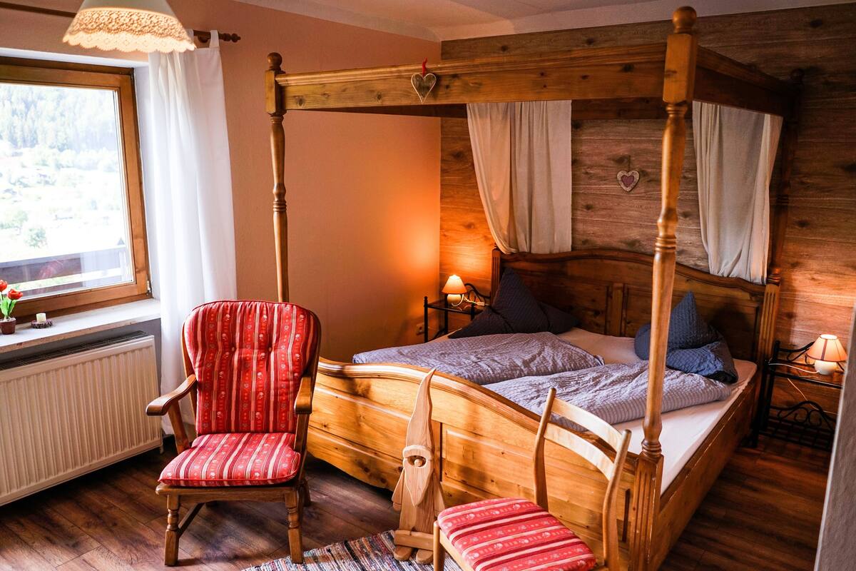 A wooden four-poster bed with a canopy, a red upholstered chair, a bedside table with a lamp, and a window with curtains.
