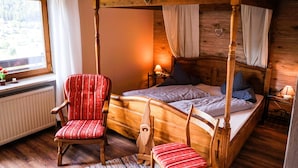 Una cama con dosel de madera, una butaca tapizada de rojo, una mesita de noche con lámpara y una ventana con cortinas.