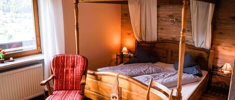 Ein hölzernes Himmelbett mit Baldachin, ein rot gepolsterter Sessel, ein Nachttisch mit Lampe und ein Fenster mit Vorhängen.