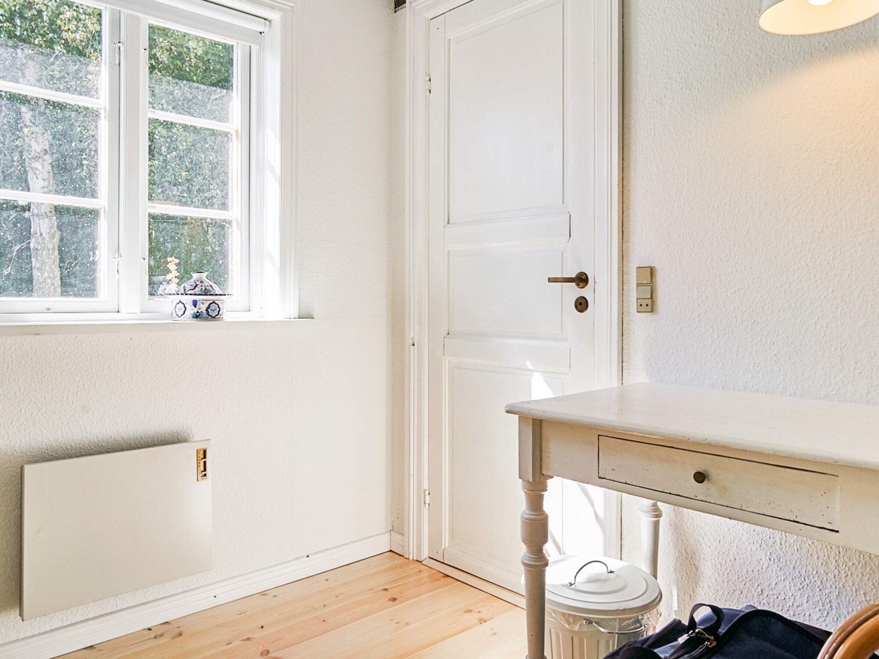 A room with a white radiator, a window with a view of trees, a white door, and a white table with a drawer.