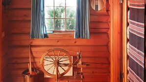 Interior de una cabaña de troncos con un rueca, una ventana con cortinas y una manta escocesa en la pared.