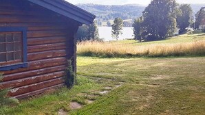 Una cabaña rústica de madera con una ventana, rodeada por un campo cubierto de hierba y un lago al fondo.