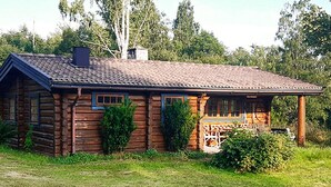 Una cabaña rústica de troncos con un porche y chimenea, rodeada de vegetación.