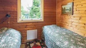 A room with wooden walls, a window with a view of greenery, two beds with floral bedding, a wall-mounted lamp, and a radiator.