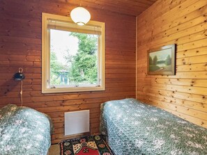 A room with wooden walls, a window with a view of greenery, two beds with floral bedding, a wall-mounted lamp, and a radiator.