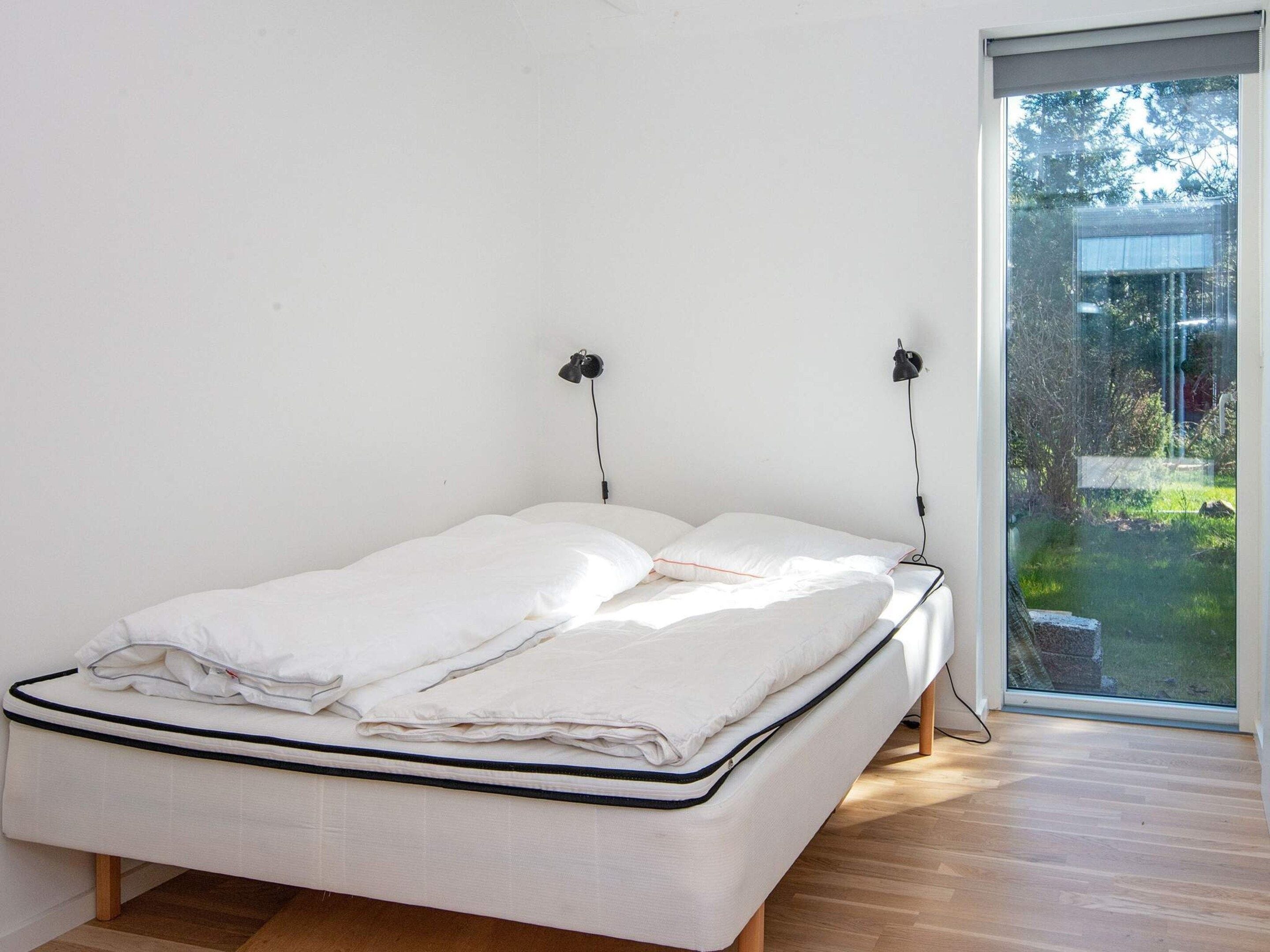 A single bed with a white mattress and pillows, a bedside table, and a window with a view of greenery outside.