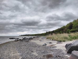 A rocky shoreline with pebbles and boulders, a forested area, and a cloudy sky.