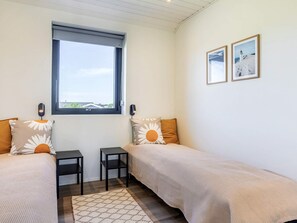 Un dormitorio con dos camas individuales, una ventana, una mesita pequeña, dos luces empotradas en la pared y dos cuadros enmarcados.