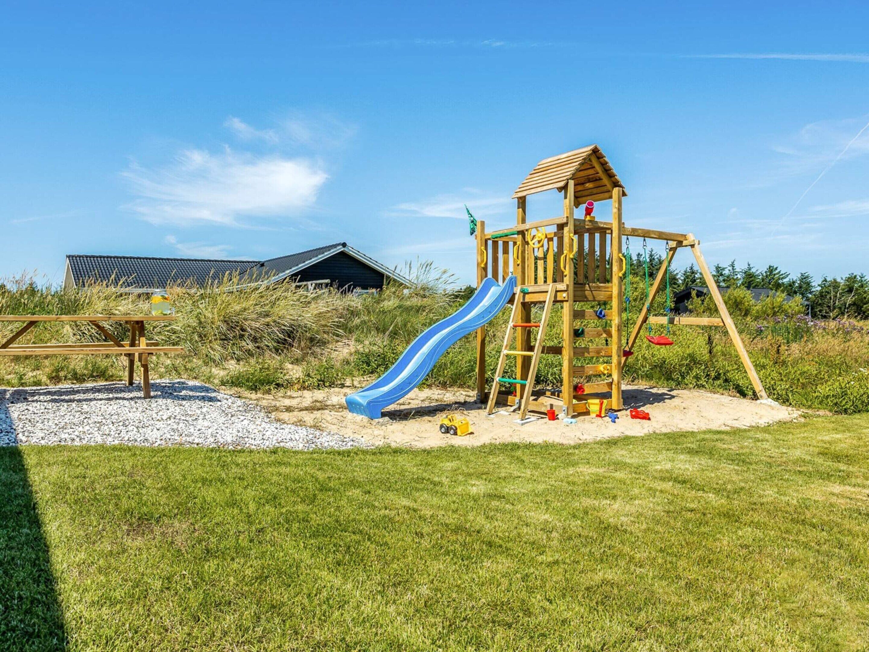 A wooden play structure with a slide and climbing elements, a picnic table, and a building with a sloped roof in the background.