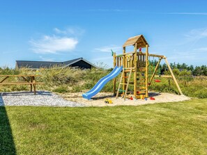 A wooden play structure with a slide and climbing elements, a picnic table, and a building with a sloped roof in the background. - 5 Star Holiday Home in Hirtshals (Hirtshals)