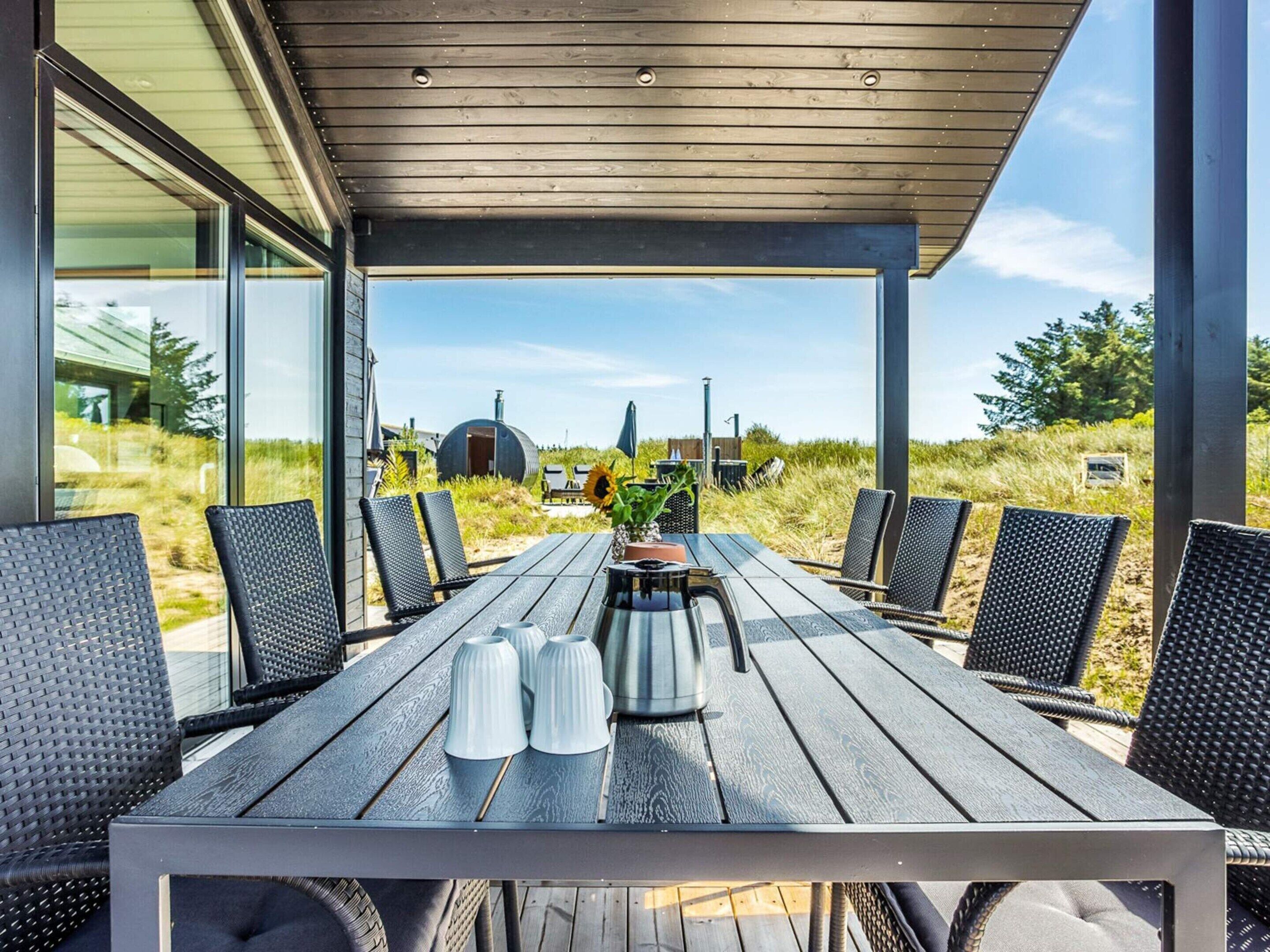 Un área de comedor al aire libre con mesa y sillas de madera, vista a la playa y un edificio de fondo.