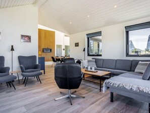 A modern living room with a grey sofa, armchairs, and a coffee table. There is a dining area in the background with a wooden accent wall and a TV mounted above it. - 5 Star Holiday Home in Hirtshals (Hirtshals)
