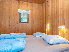 A wooden room with a bed, two pillows, a window with blinds, and a wall-mounted lamp.