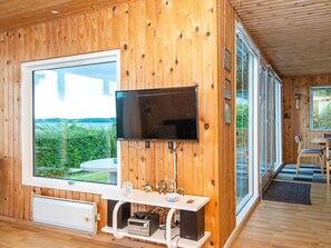 A wooden-paneled room with a large window, a flat-screen TV mounted on the wall, a small table with a chair, and a heater on the floor.