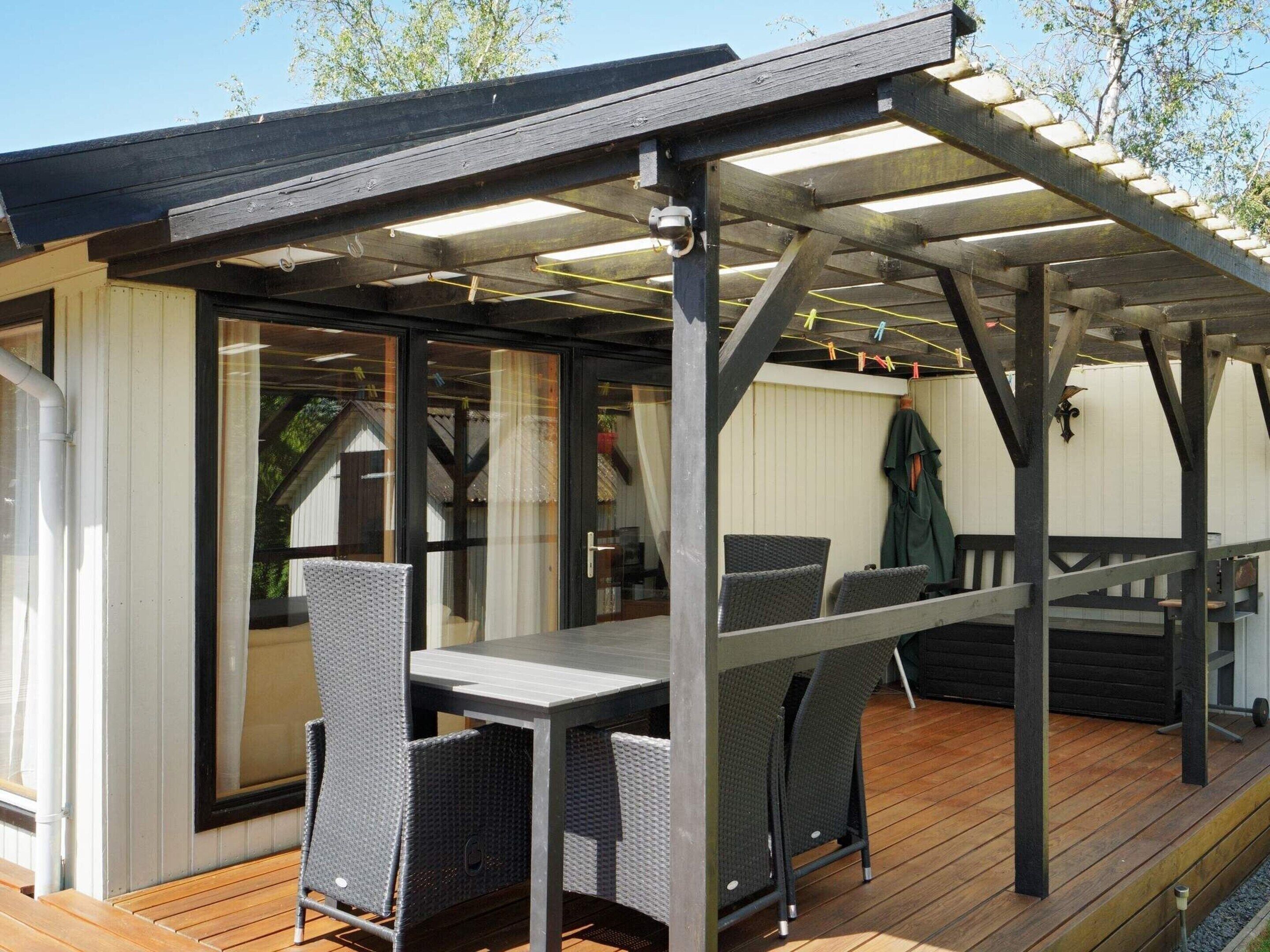 A covered outdoor seating area with a table and chairs, wooden deck flooring, and a pergola structure.