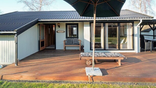 A covered outdoor seating area with a bench and umbrella, a wooden deck, and a building with a metal roof and sliding glass doors. - 5 Person Holiday Home in Follenslev (Follenslev)