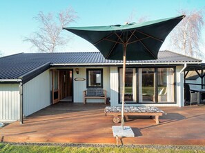 Et overdækket udendørs område med bænk og parasol, en træterrasse og en bygning med ståltag og skydedøre.