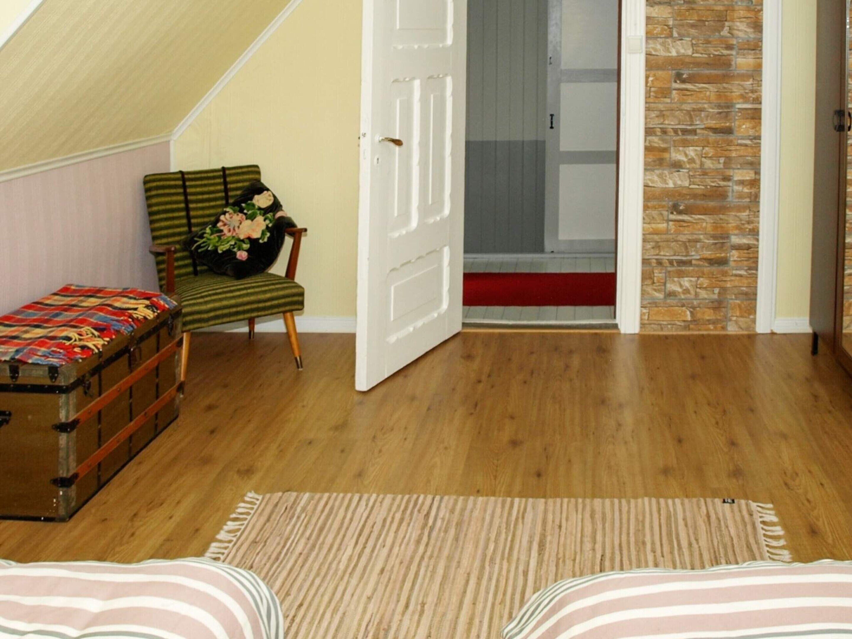 A room with a wooden floor, a bench with a colorful blanket, a chair with a floral cushion, and a door leading to a hallway with a red carpet.