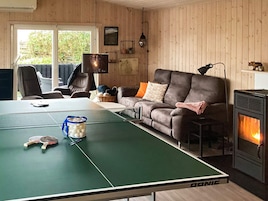 A room with a ping pong table, a sofa, a fireplace, and a window with a view of greenery.
