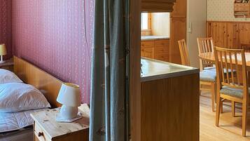 A room with a bed, a nightstand with a lamp, a wooden table with chairs, and a kitchen area with wooden cabinets and a refrigerator.