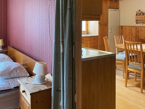 Una habitación con cama, mesita de noche con lámpara, mesa de madera con sillas y una zona de cocina con armarios de madera y un refrigerador.