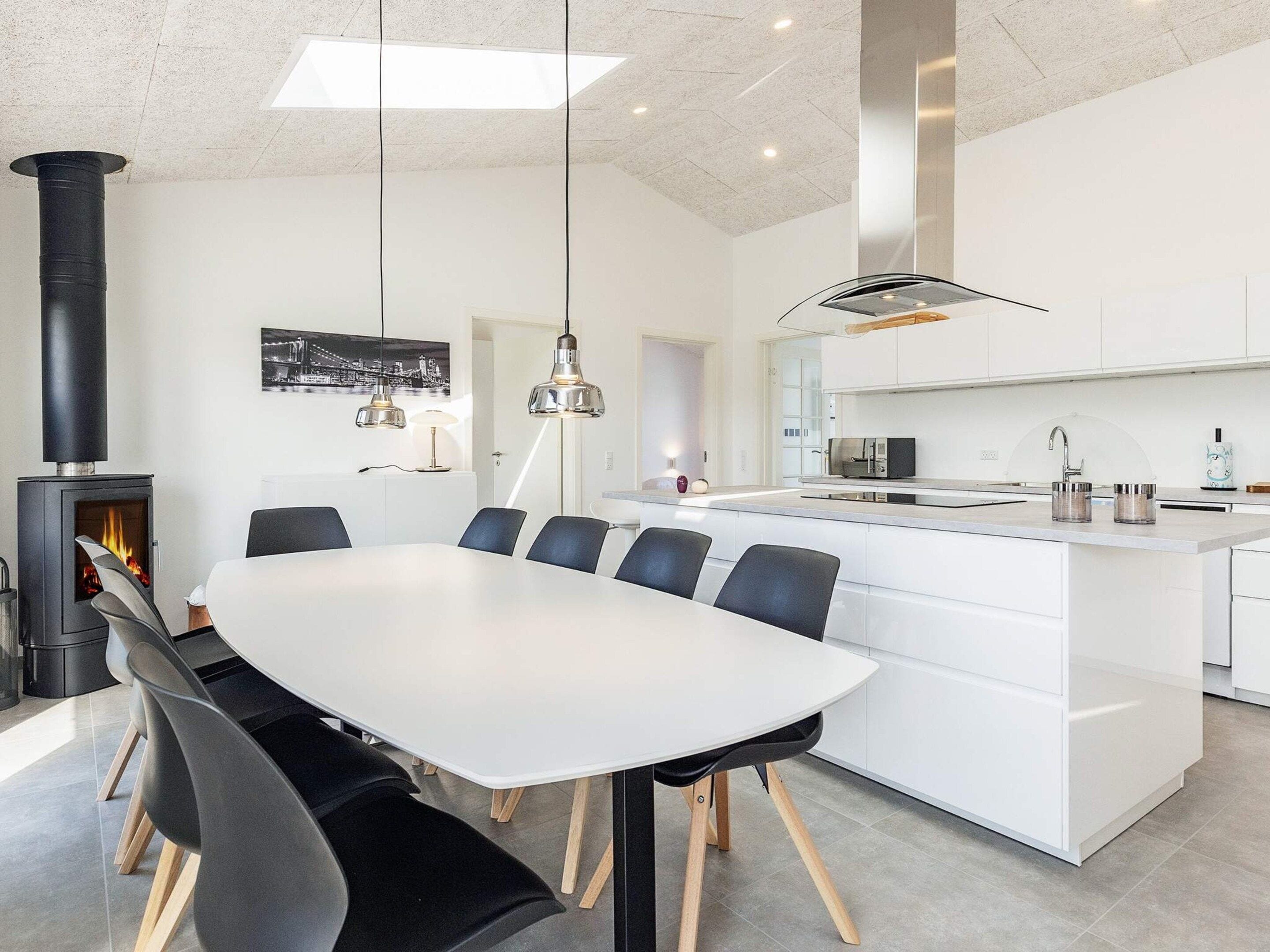 A modern kitchen with a central island, a dining area with a white table and chairs, and a black wood-burning stove.