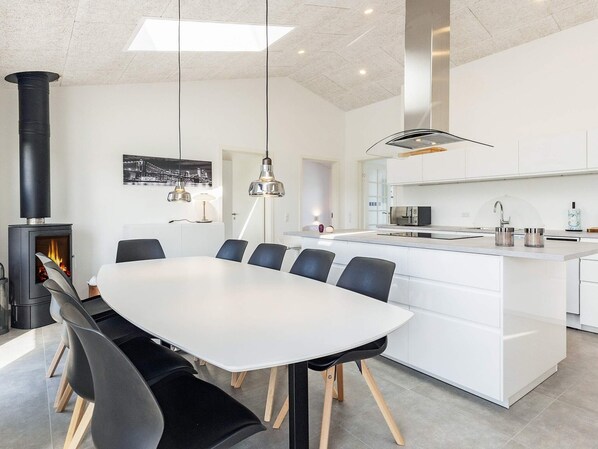 A modern kitchen with a central island, a dining area with a white table and chairs, and a black wood-burning stove.