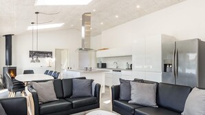 A modern living room with black leather sofas, a white dining table, and a kitchen area with white cabinets and stainless steel appliances. - 10 Person Holiday Home in Lokken (Lokken)